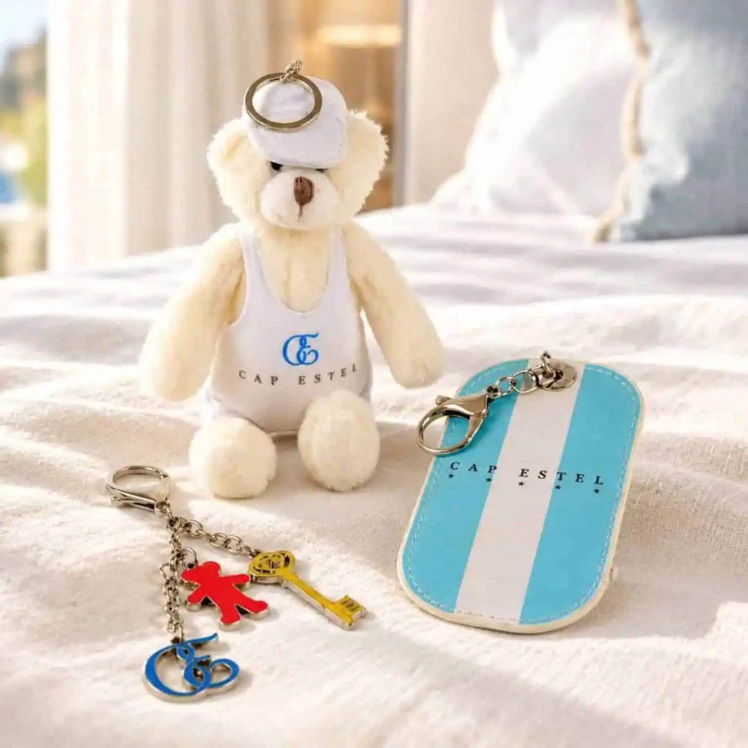 Ours en peluche blanc avec logo Cap Estel assis sur un lit, accompagné de porte-clés colorés et d'une pochette rayée bleu et blanc