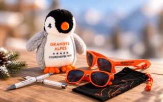 Goodies Grandes Alpes Courchevel Peluche manchot aux yeux orange vif, stylos et lunettes de soleil orange marqués 'Grandes Alpes Courchevel 1850' posés sur une surface en bois
