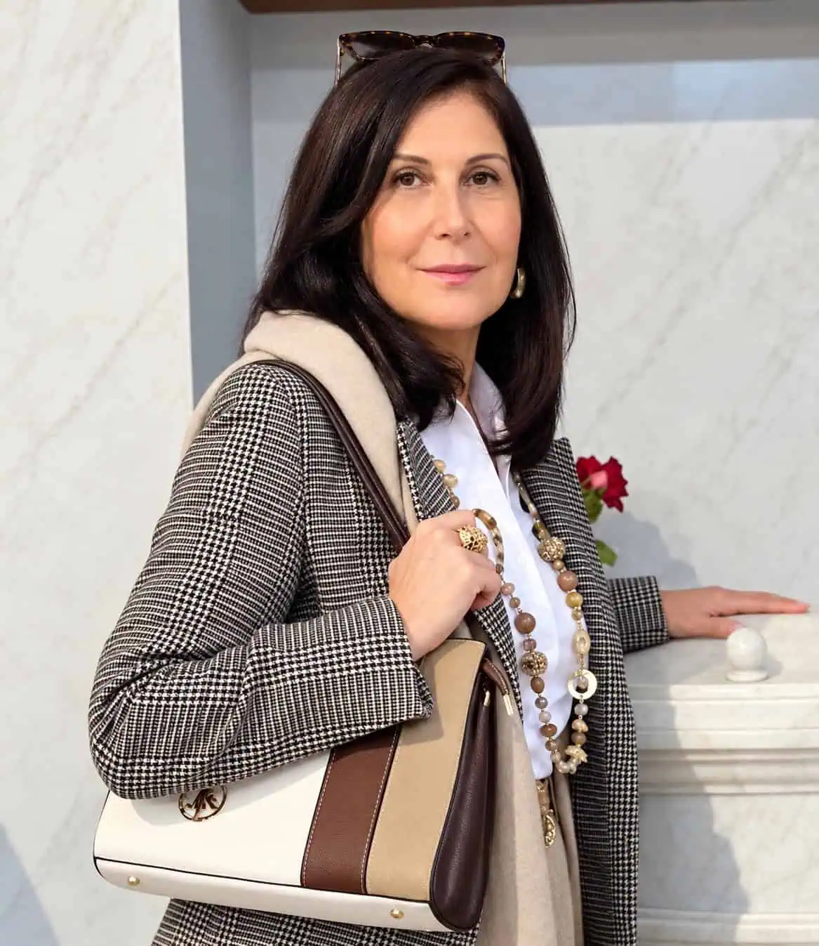 Sylvie Delsanti Femme portant un blazer à carreaux noir et blanc avec un col crème, tenant un sac à main beige et marron, souriante.