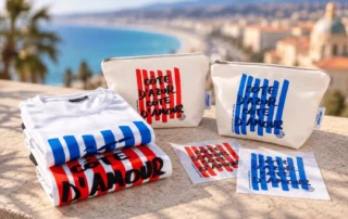 Gamme textile rayée « Latitude » Collection de vêtements et accessoires avec motif rayé bleu et rouge, présentés sur une terrasse avec vue panoramique sur une baie méditerranéenne.