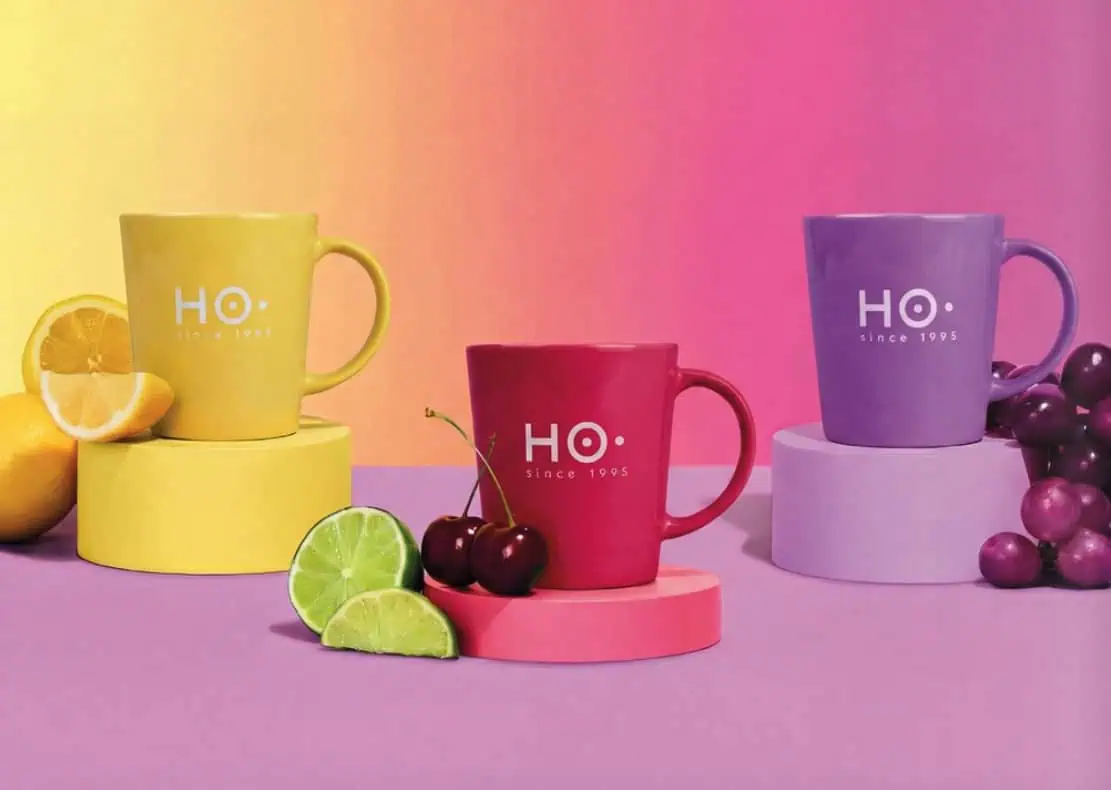 Trois mugs colorés HO de la collection depuis 1995, présentés sur des socles assortis avec des fruits frais comme accessoires de mise en scène